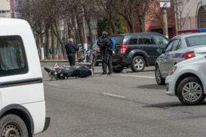 Motorcycle rider lies beside fallen bike in street as officer and bystanders assess the scene near parked cars.