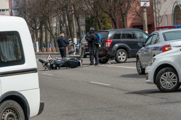AccidentInTheCityABrokenMotorcycleAndACar - Grooms Law Firm Motorcycle rider lies beside fallen bike in street as officer and bystanders assess the scene near parked cars.