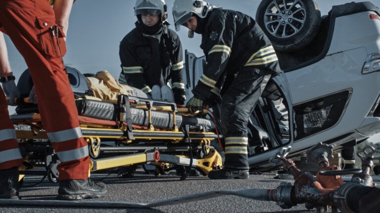 OnCarCrashTrafficAccidentSceneRescueTeamOfFirefighters - Grooms Law Firm Emergency responders in gear prepare a stretcher beside a flipped car on a roadway during a rescue.1/5