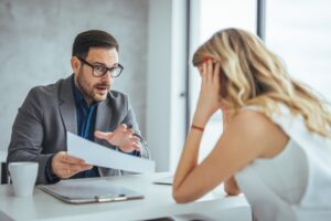 AngryBusinesswomanDissatisfiedByContractConditionsArguingWithBusinessPartner - Grooms Law Firm Businessman in glasses explains a document to a stressed woman across a desk in a bright office setting.