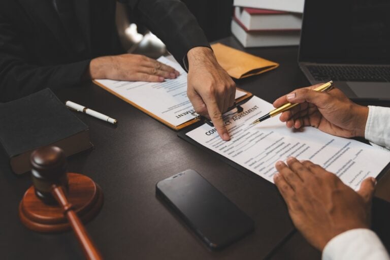 TheLawyerIsReadingTheStatuteOfLimitationsConsultingWith - Grooms Law Firm Two people sign a contract on a dark desk, with a gold pen, gavel, smartphone, and laptop nearby.