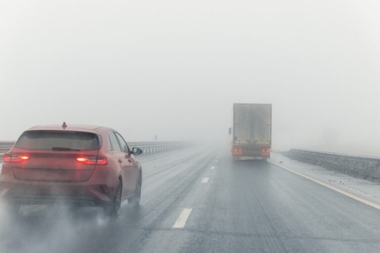 DriverPovOnTrafficOnBlueFoggyMistyRainySlush - Grooms Law Firm Red SUV ahead on a foggy highway with a large truck in the distance and wet road conditions reducing visibility.
