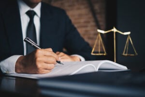 Person in a suit signs documents at a desk, with the scales of justice visible in the background.