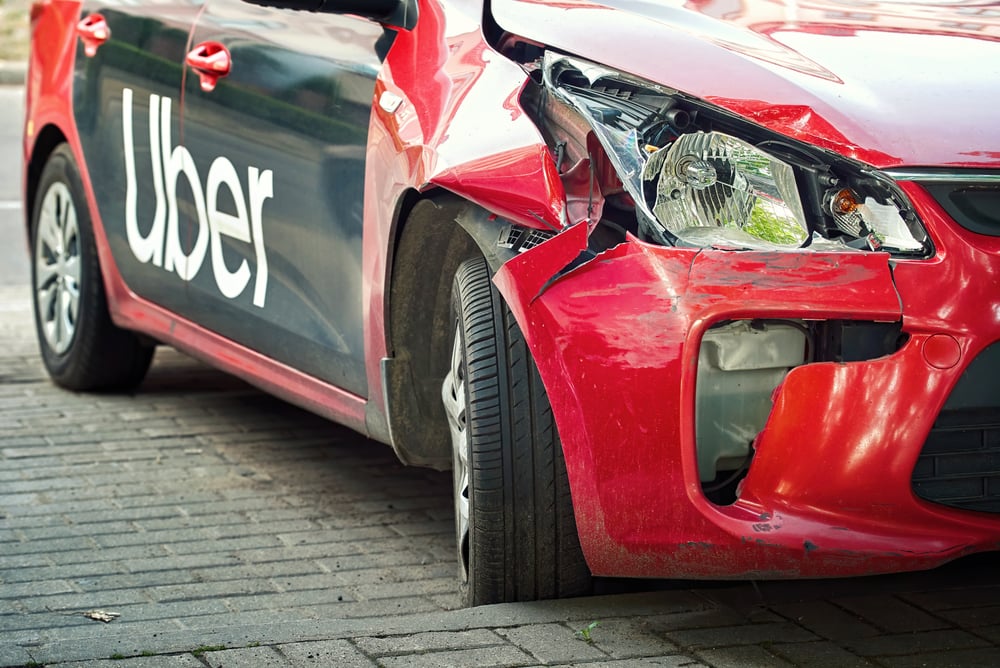 Red Uber car with severe front-end damage from a collision, showing a crumpled bumper and exposed headlight area on a street.
