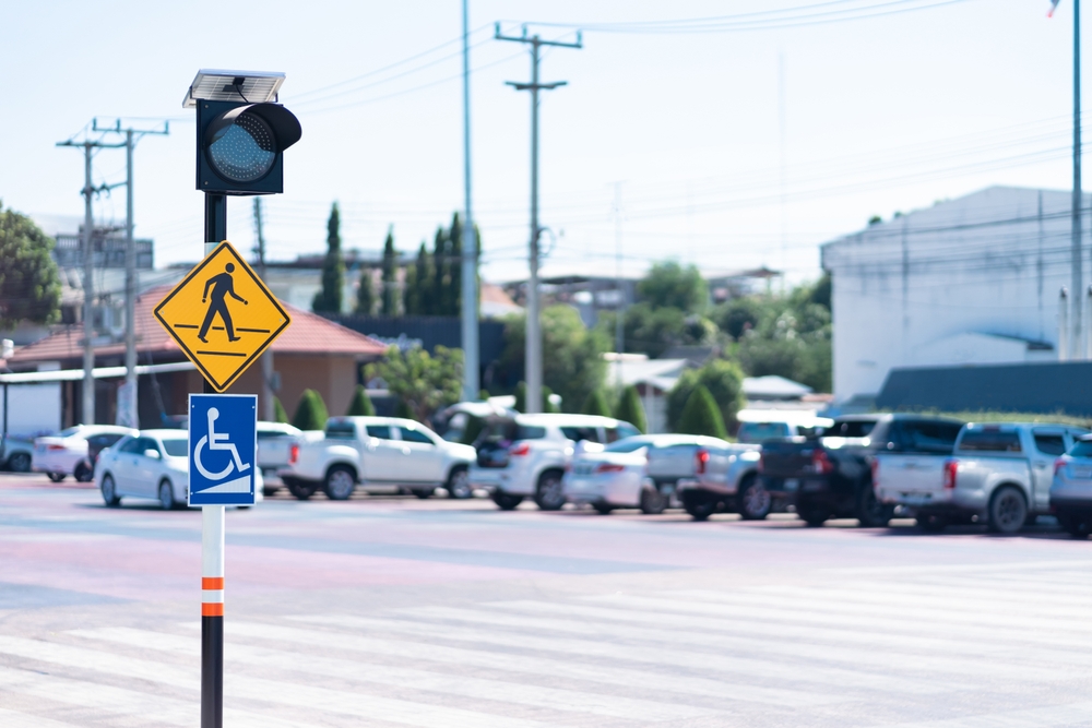 Pedestrian crossing warning sign and blue wheelchair accessibility sign on a pole at a busy crosswalk with cars in the background, urban scene.
