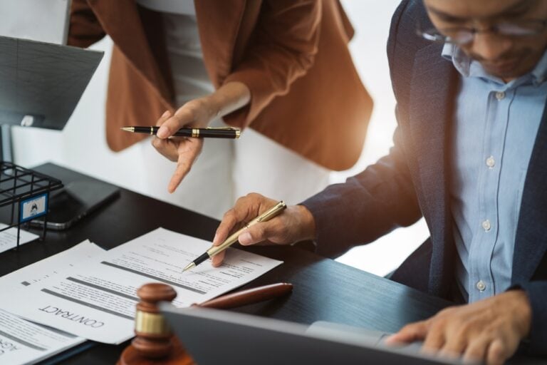 Two professionals sign a contract at a desk, with one person pointing a pen while the other writes with a gold pen; a gavel sits nearby.