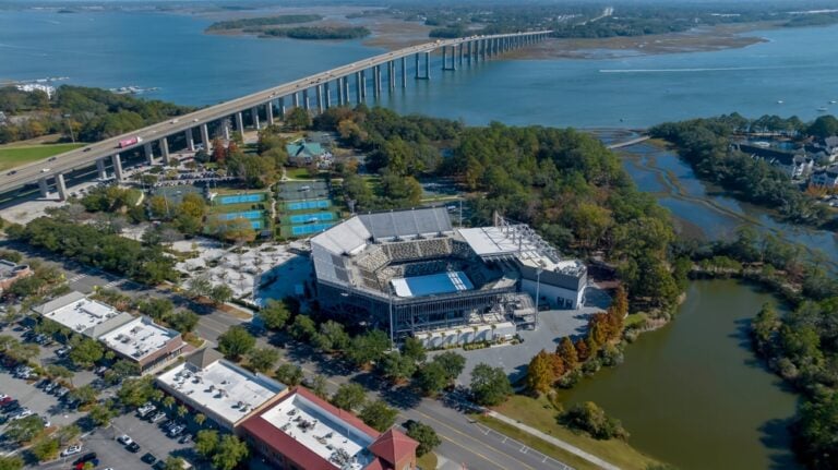 Aerial view of a large stadium surrounded by trees, with a long bridge crossing the water in the background. Nearby tennis courts and parking areas are visible.