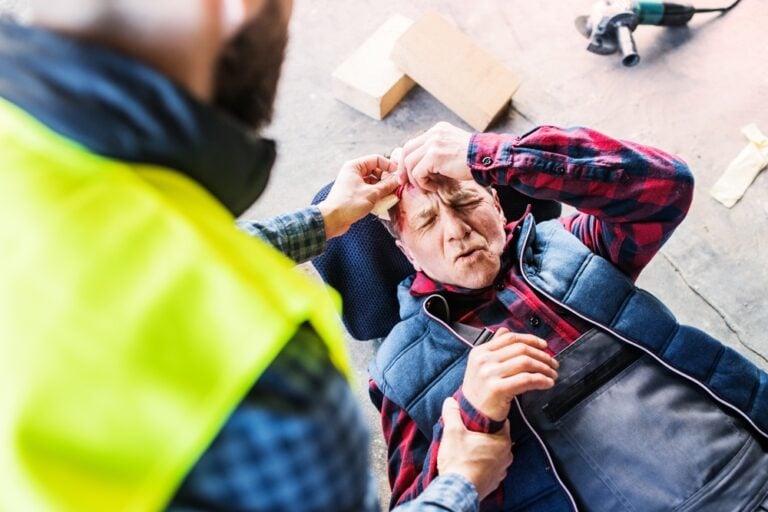 ColleagueCallingAmbulanceAndGivingFirstAidToInjuredWorker - Grooms Law Firm Man lying on a workshop floor clutching his head as a colleague in a yellow safety vest checks him, with tools and bricks nearby.