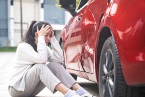 Woman sitting on the ground beside a red car with a dented door, hands on her head in distress after a crash.