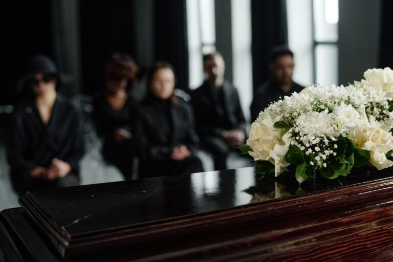 Polished dark coffin with a white floral arrangement on top, mourners in black seated in the background at a funeral.
