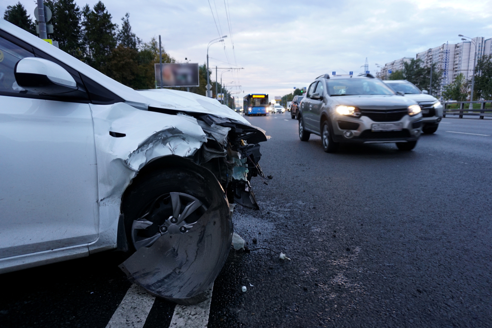 A white car with severe front-end damage stopped at the roadside as traffic passes on the highway