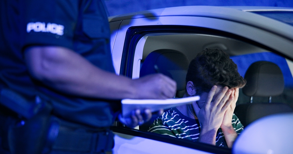 A police officer writing a citation at the window of a car at night while the driver sits with his head in his hands