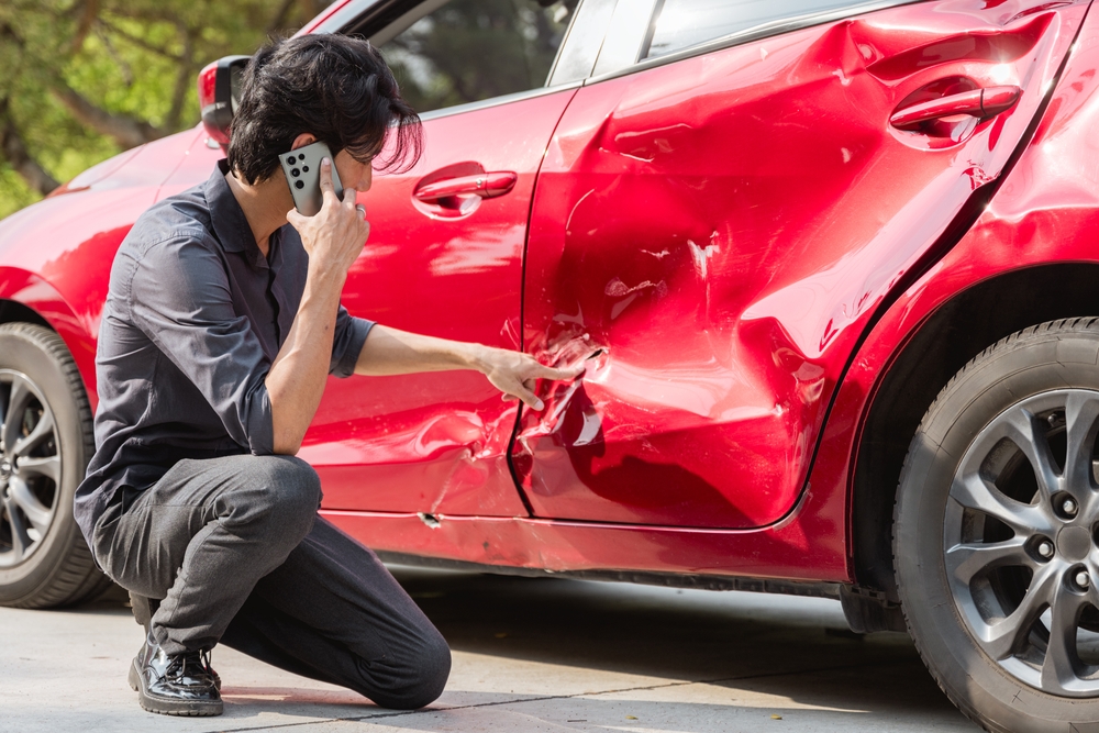 A man kneeling beside his red car, pointing at a deep dent in the door while speaking on his phone after a collision